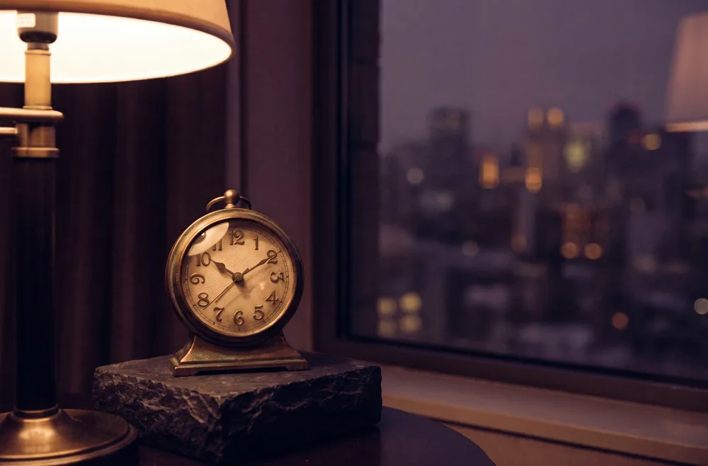 A small vintage brass table clock on dark stone at dusk, warm lamp glow on its face.